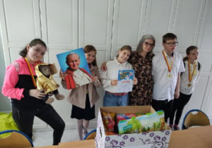 Na zdjęciu znajduje się sześć osób: pięć uczniów i nauczycielka religii. Uczniowie mają zawieszone na szyi medale. Uczennica stojąca przy oknie trzyma maskotkę - jedną z nagród, druga od lewej trzyma portret św. Jana Pawła II. Uczennica stojąca przy naucZycielce prezentuje dyplom zdobyty na Przeglądzie. Wszyscy są uśmiechnięci i szczęśliwi z powodu udziału w konkursie.