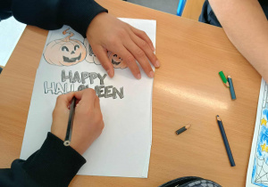 Dzieci siedzą przy stolikach i kolorują obrazek przedstawiający dynię oraz stracha na wróble w klimacie Halloween.