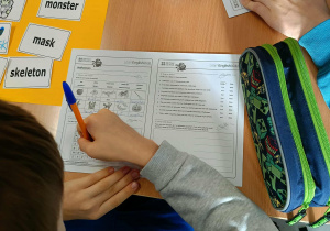 Grupa uczniów siedzi przy stoliku i pracuje nad kartami z zadaniami językowymi dotyczącymi Halloween.